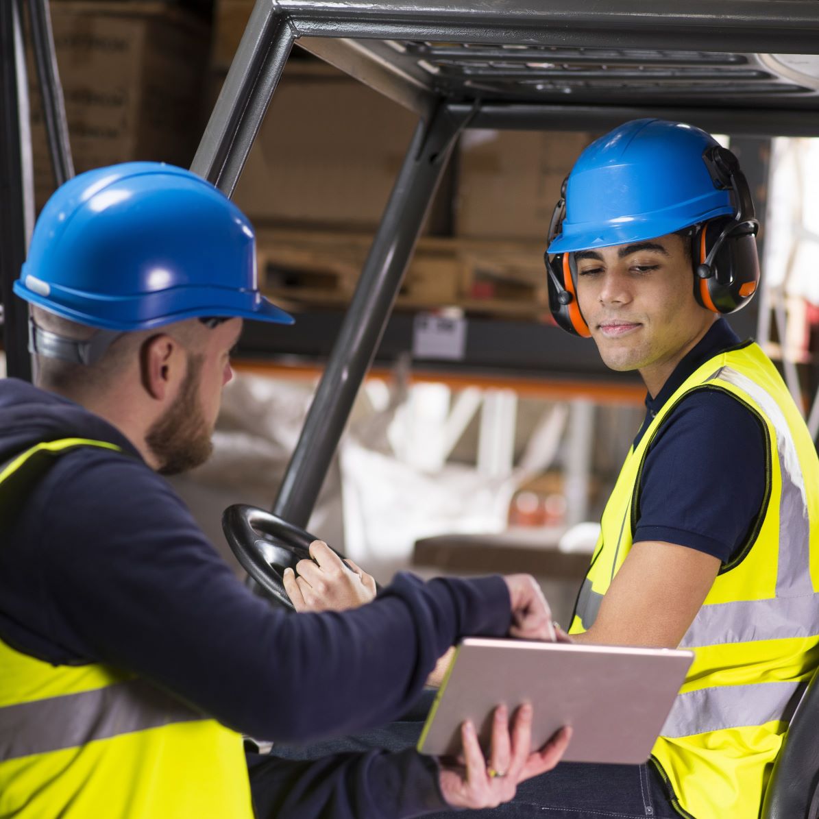 Two individuals in safety vests and helmets engaged in forklift training with a tablet in a warehouse setting.