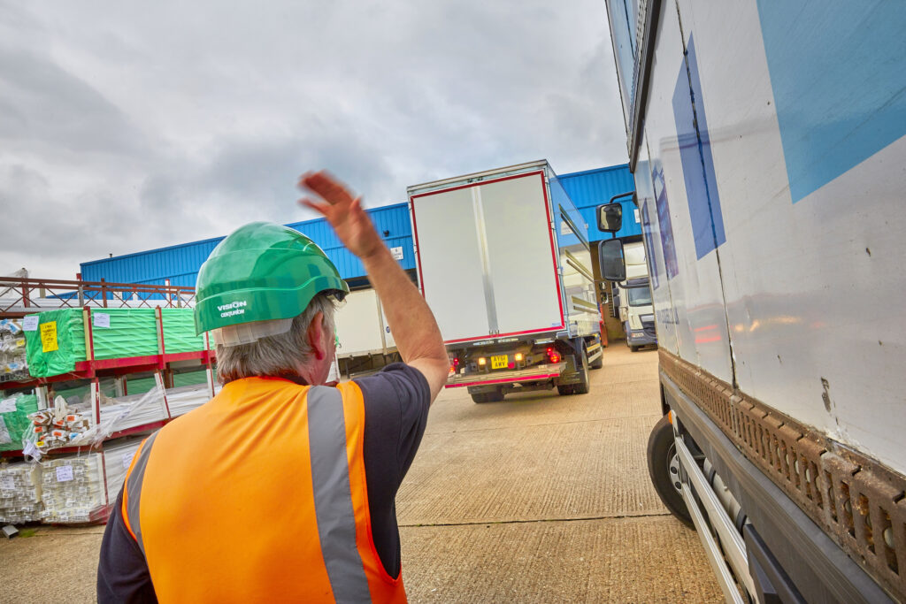 A lorry banksman signals drivers between parked trucks at an industrial yard, following lorry banksman training with Milne Safety