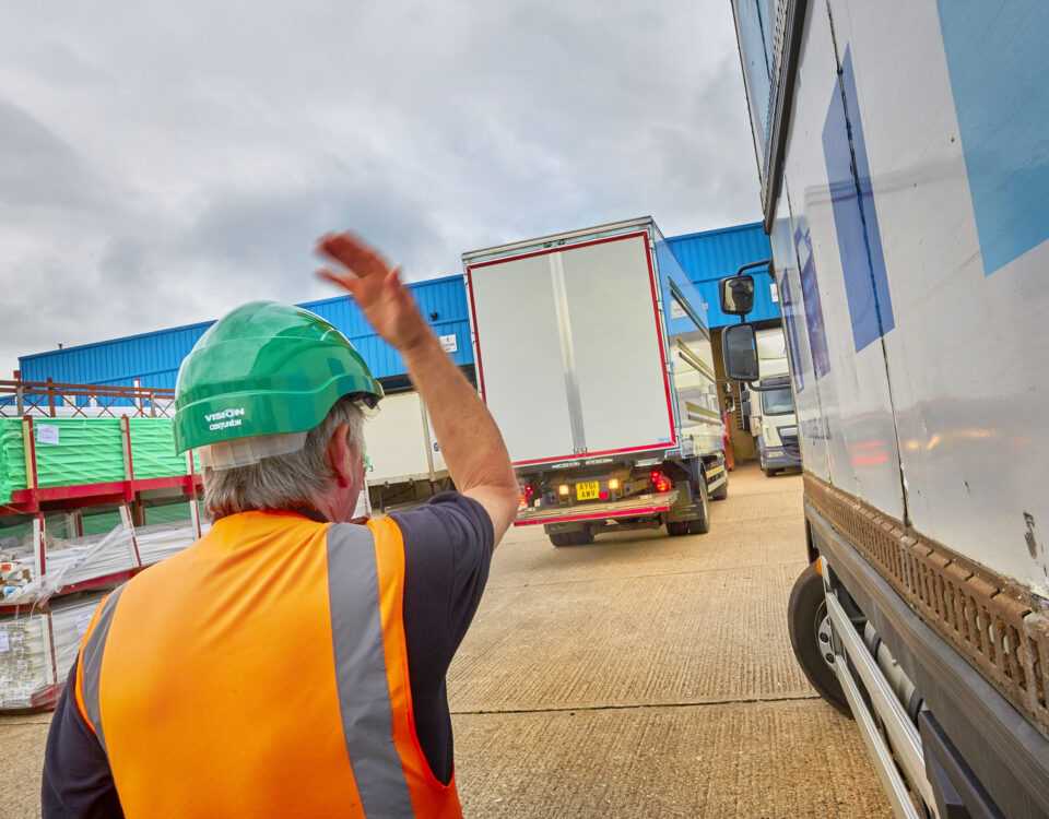 A lorry banksman signals drivers between parked trucks at an industrial yard, following lorry banksman training with Milne Safety