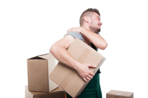 Man in a gray shirt holding a cardboard box while grimacing in discomfort, surrounded by more boxes.