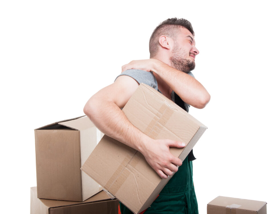 Man in a gray shirt holding a cardboard box while grimacing in discomfort, surrounded by more boxes.
