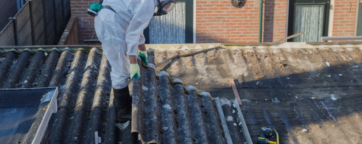 A worker in a white protective suit and respirator inspects corrugated asbestos roofing on a house rooftop.
