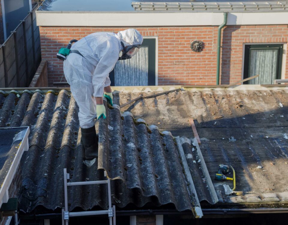 A worker in a white protective suit and respirator inspects corrugated asbestos roofing on a house rooftop.