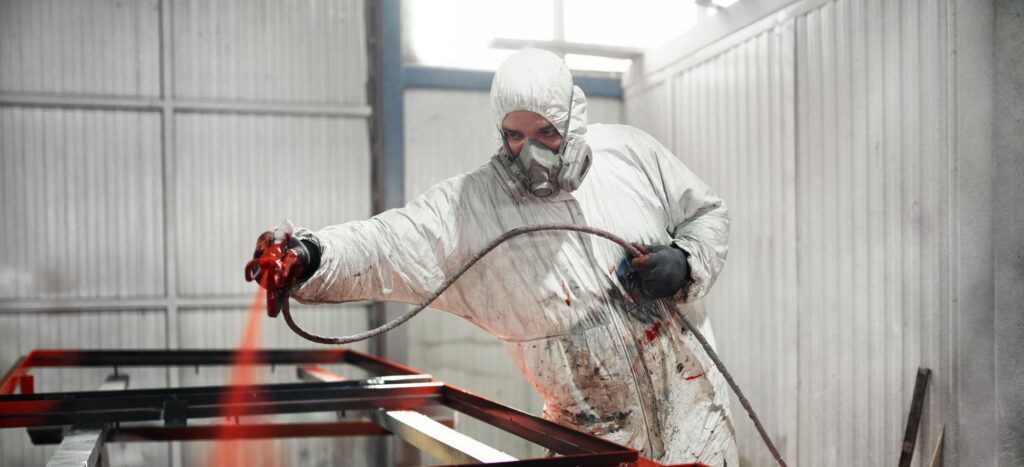 A worker in a white protective suit and respirator sprays red coating onto a metal frame inside an industrial bay. Using a face mask following a face fitting test