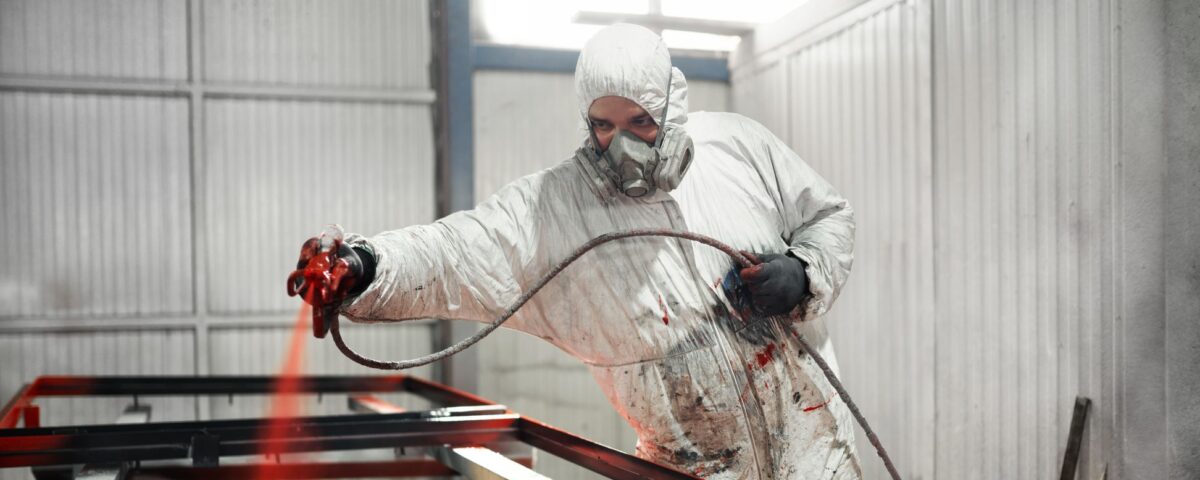 A worker in a white protective suit and respirator sprays red coating onto a metal frame inside an industrial bay. Using a face mask following a face fitting test