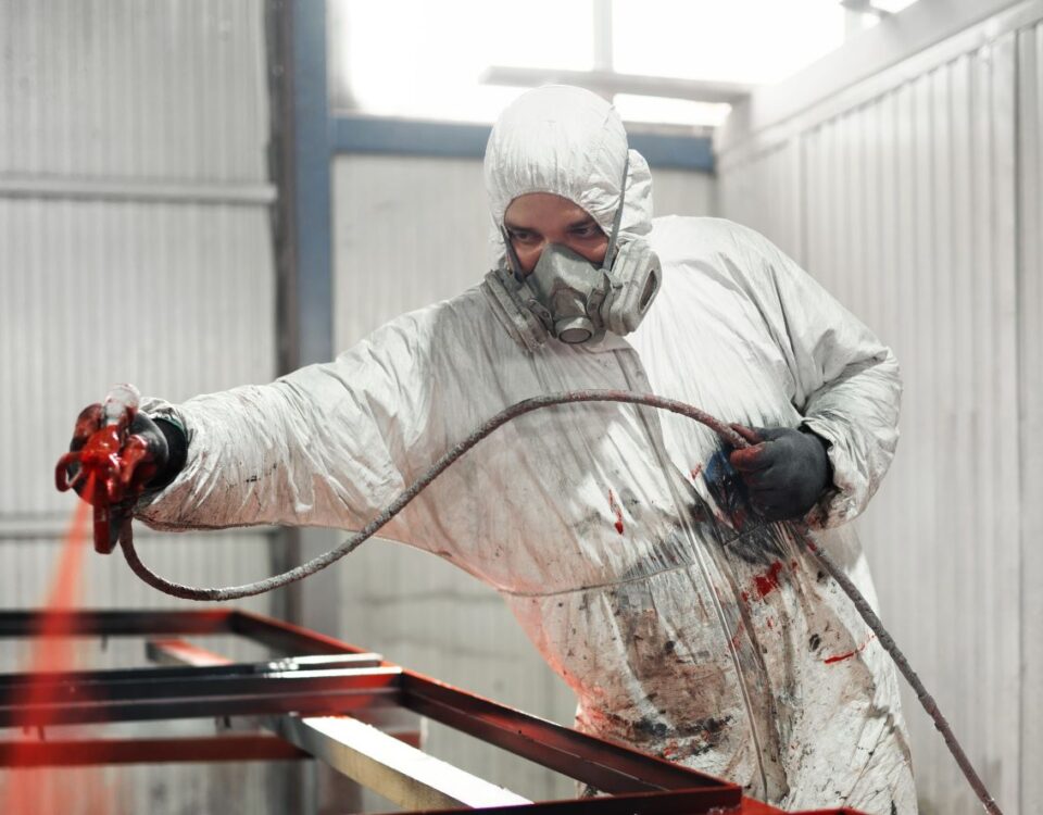 A worker in a white protective suit and respirator sprays red coating onto a metal frame inside an industrial bay. Using a face mask following a face fitting test
