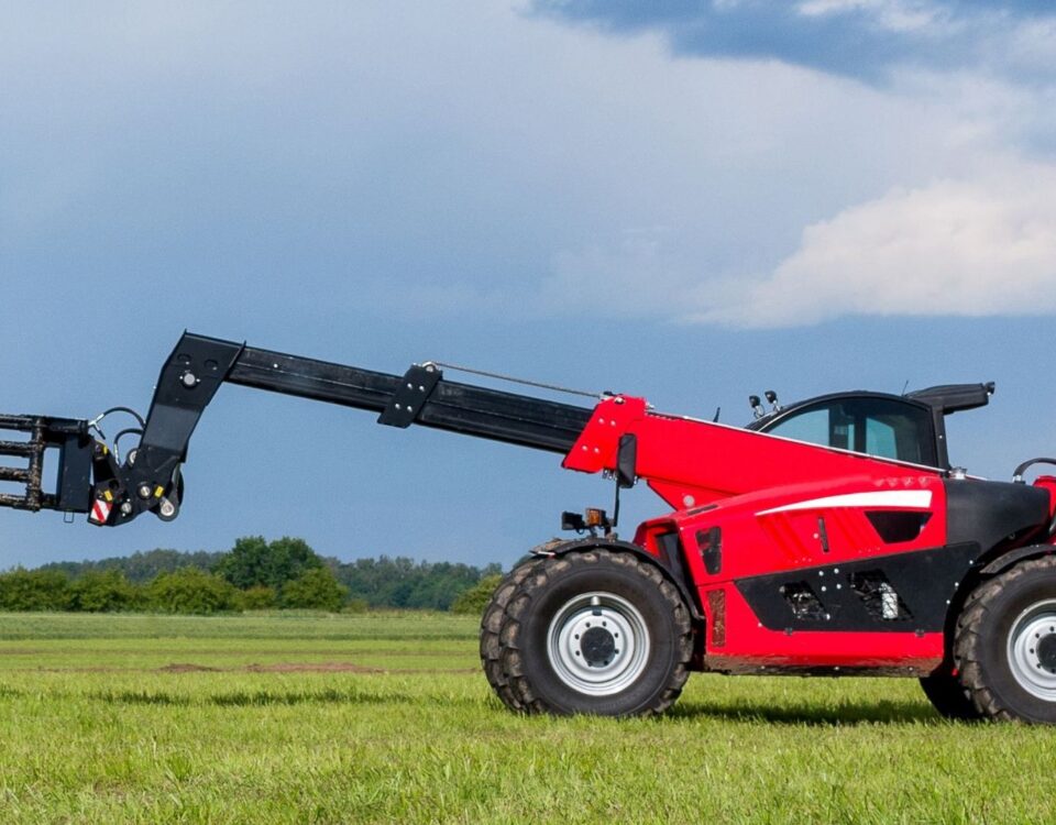 A red telehandler rests on grass with its boom extended leftward beneath a dramatic cloudy sky.