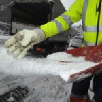 Worker in a high-visibility jacket spreading salt from a shovel on a snowy road surface.