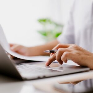 Online training session with laptop and pen in a bright office Hands use a laptop trackpad while holding a pen, with paperwork spread out during an online training session.