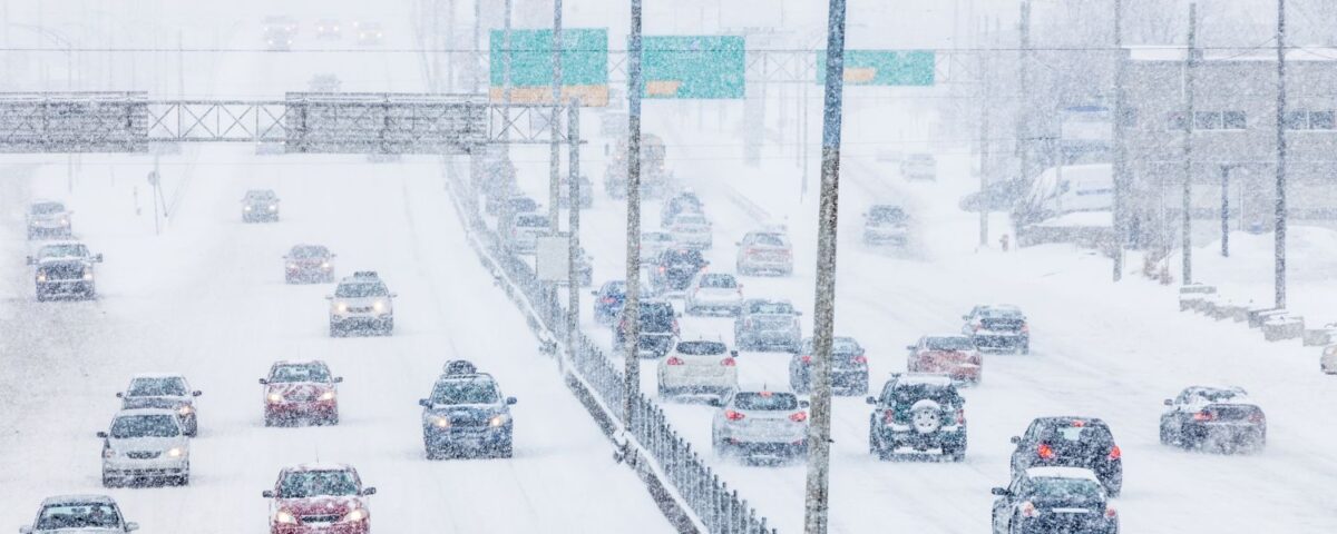 Vehicles traveling on a snow-covered highway during a winter snowstorm with reduced visibility.