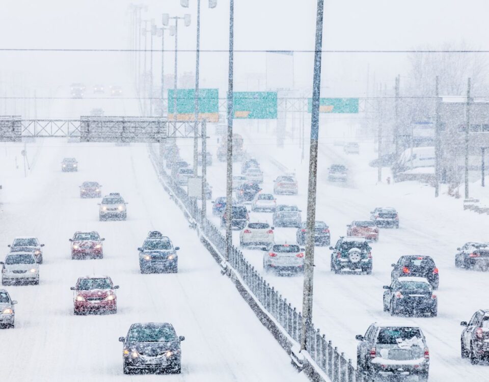 Vehicles traveling on a snow-covered highway during a winter snowstorm with reduced visibility.
