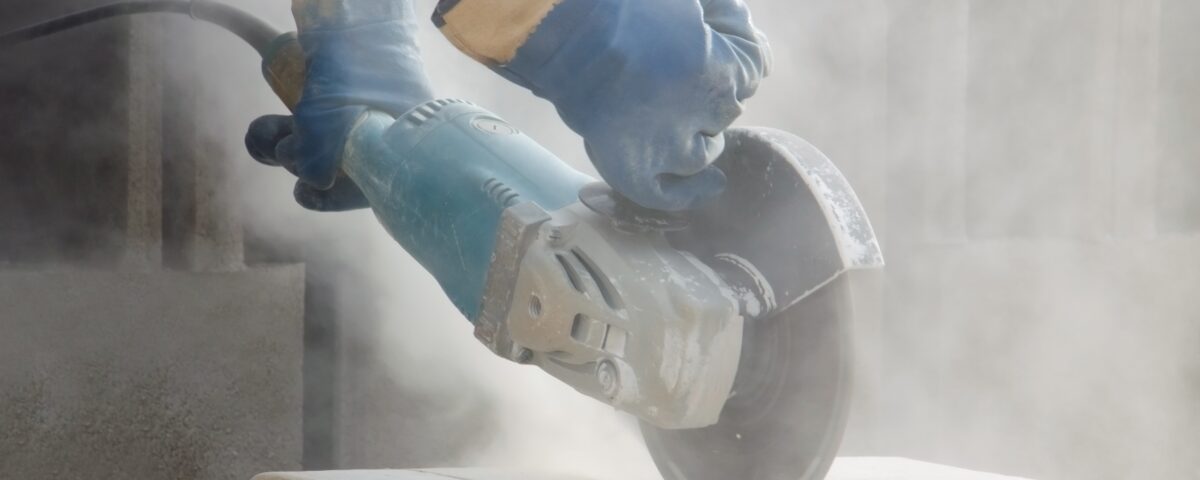 Blue-gloved hands cut a light stone block with an angle grinder, sending fine dust into the air during masonry work.