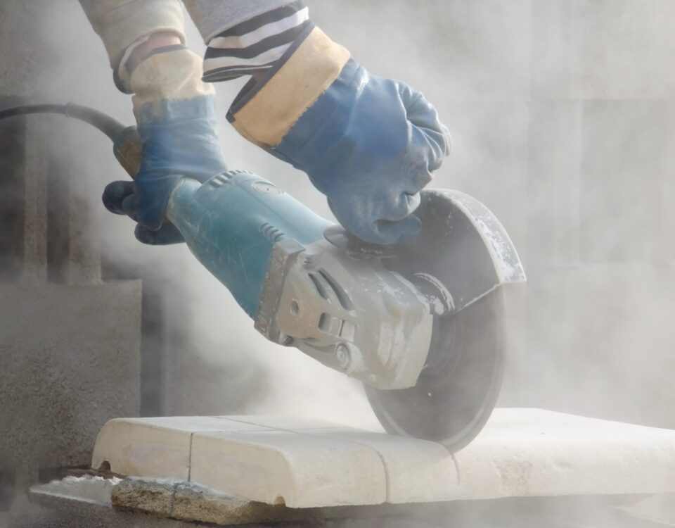 Blue-gloved hands cut a light stone block with an angle grinder, sending fine dust into the air during masonry work.