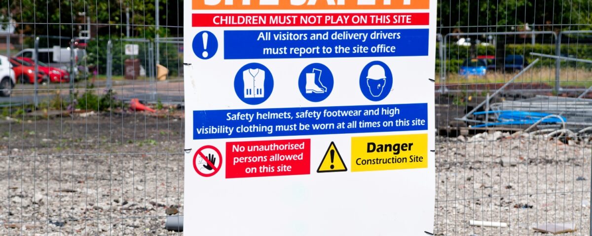 A site safety notice on a fenced construction area lists rules for visitors, helmets, footwear and high-visibility clothing.