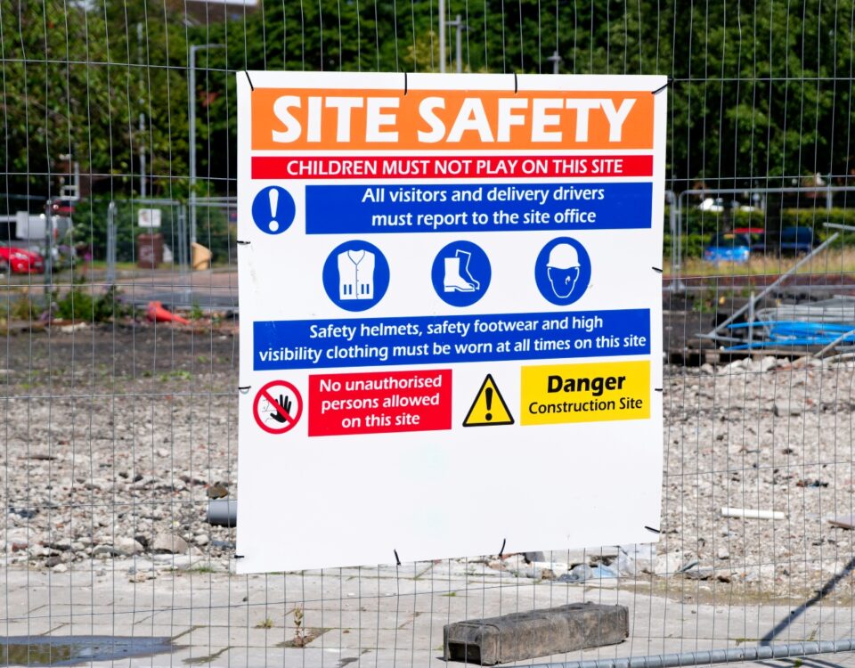 A site safety notice on a fenced construction area lists rules for visitors, helmets, footwear and high-visibility clothing.