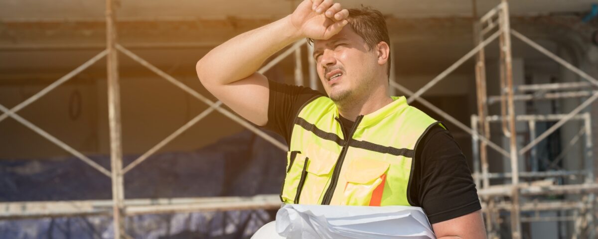 A construction worker in a high-visibility vest shields his face from the sun while holding a hard hat at a scaffolding site.