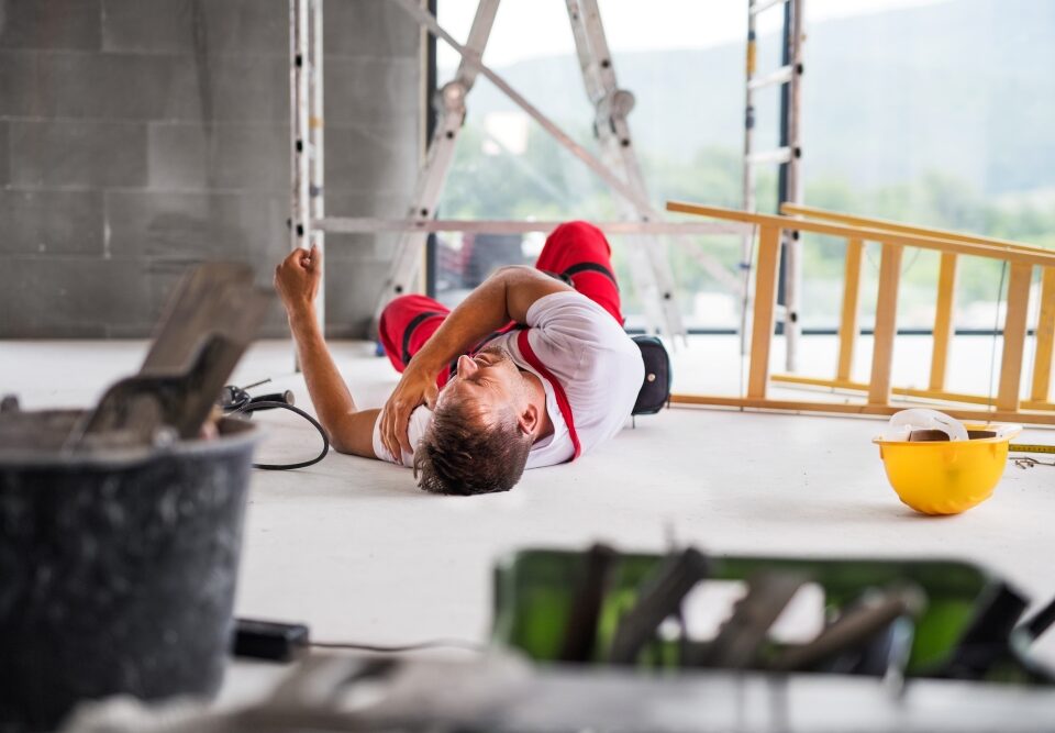 A worker in red overalls lies on the floor after a fall, with a yellow hard hat, ladder, and tools nearby.