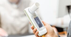 A hand holds a white lung function meter with a small screen, while a blurred person sits in the background of a clinic room.