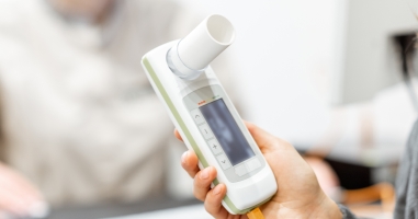 A hand holds a white lung function meter with a small screen, while a blurred person sits in the background of a clinic room.