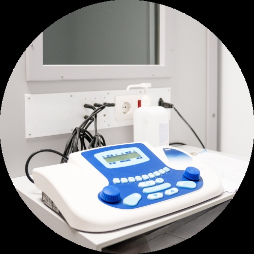 A compact audiometric testing unit sits on a clinic counter beside tangled cables, a wall outlet, and a white bottle.