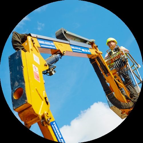 A worker in a yellow hard hat operates a yellow MEWP boom lift while standing high against a clear blue sky.