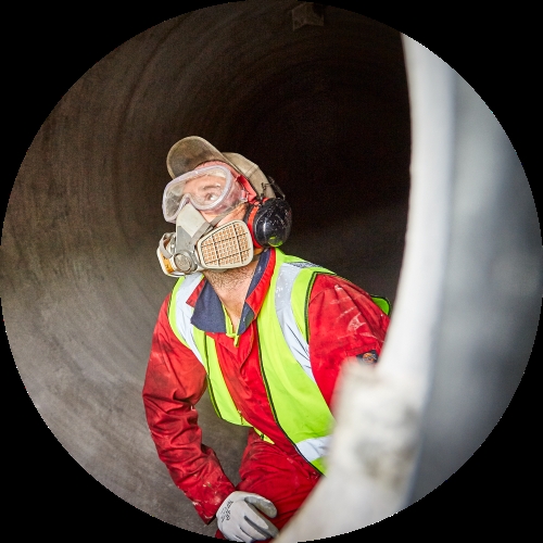 A worker in red coveralls and a respirator looks upward inside a large cylindrical confined space.