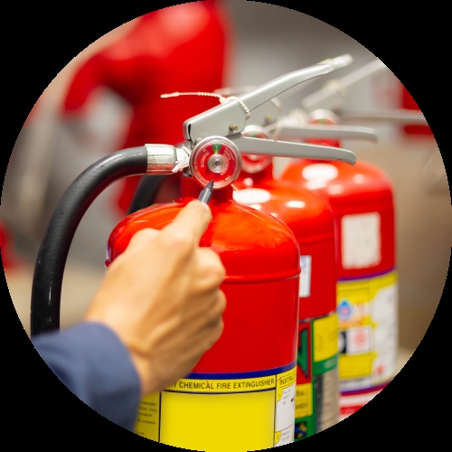 A hand checks the pressure gauge on a red chemical fire extinguisher, with more extinguishers lined up behind it.