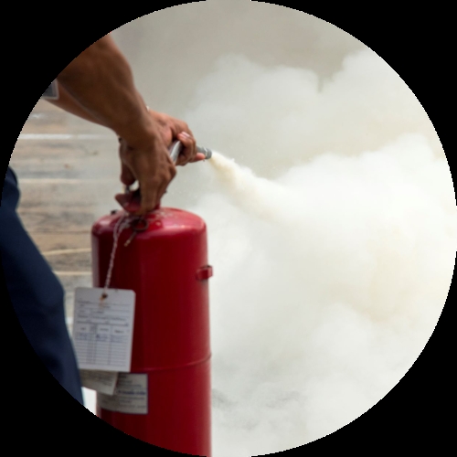 A person grips a red fire extinguisher and sprays foam into the air as thick white smoke or vapour spreads across the frame.