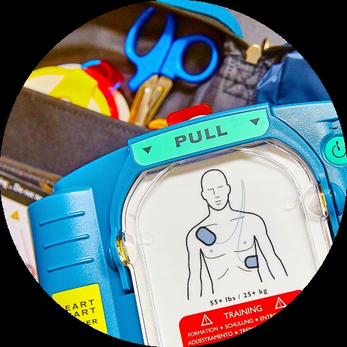 A blue AED trainer shows a pull tab, training pads, and a chest diagram beside scissors and safety gear.