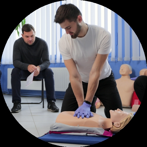 A man in purple gloves performs chest compressions on a CPR mannequin while another attendee observes from a chair nearby.