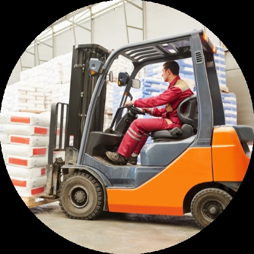 A forklift operator in red workwear drives an orange lift truck past stacked pallet loads inside a warehouse.