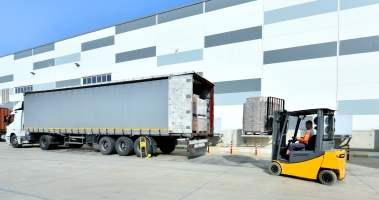 A yellow forklift driven by a worker in an orange vest moves pallets beside a long grey trailer outside a modern warehouse.