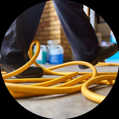 A worker's boots step past a yellow cable stretched across the floor, creating a clear trip hazard in a workshop.