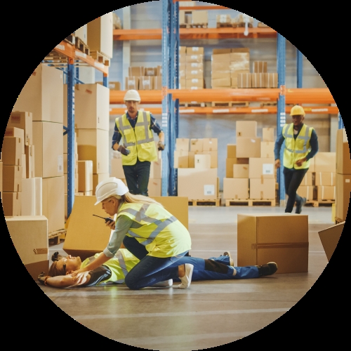 Warehouse workers in hi-vis vests and hard hats assist an injured colleague on the floor beside stacked cardboard boxes.