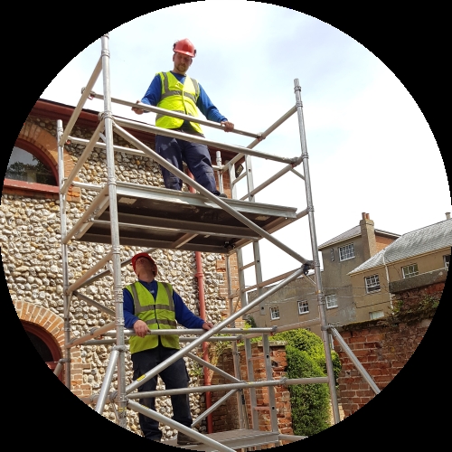 Two workers in hard hats and hi-vis vests stand on metal tower scaffolding beside a brick and flint building.