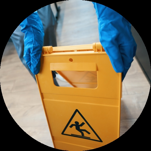 Blue-gloved hands hold a yellow folding warning sign marked with a slipping hazard symbol on a corridor floor.