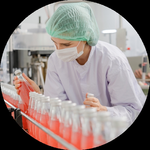 A masked factory worker in a green hairnet checks red bottled drinks moving along a production line.