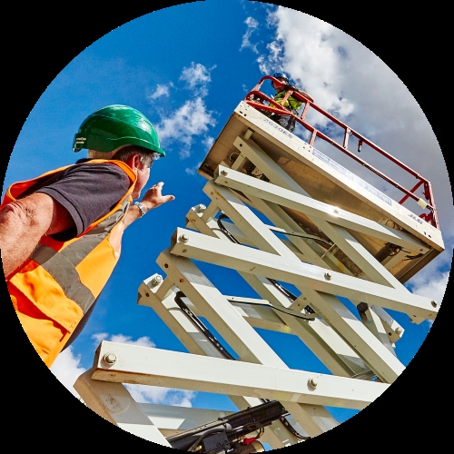A worker in a green hard hat and orange safety vest gestures toward a raised scissor lift against a bright sky.