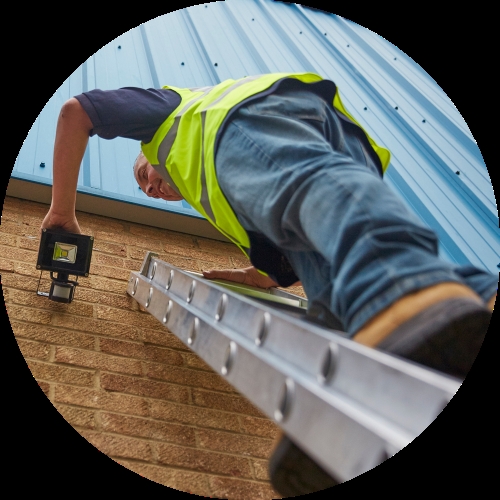 A worker in a high-visibility vest climbs a ladder beside a brick wall and blue metal cladding while holding a work light.