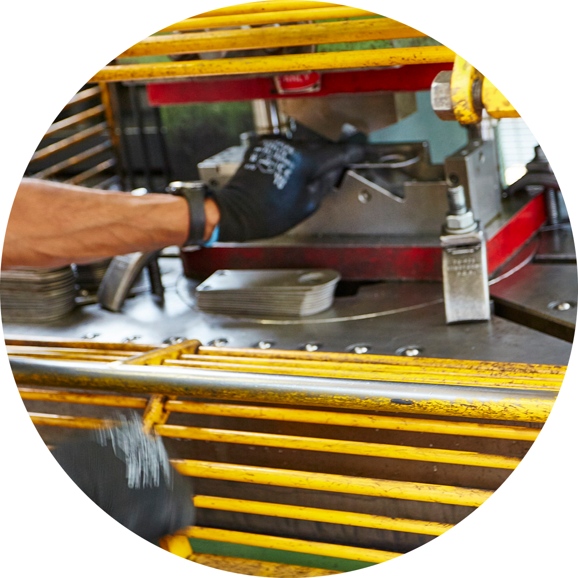 A gloved worker reaches into an industrial power press while stacked metal parts rest on the machine bed.